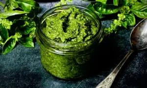 Ein Glas mit selbstgemachtem Pesto, umgeben von frischem Basilikum. Daneben liegt ein Löffel.