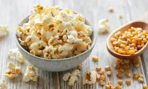 Schüssel mit frisch gemachtem Popcorn, daneben eine Holzschaufel mit Maiskörnern. So einfach ist Popcorn selber machen!
