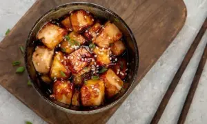 Ofen-gebackener Tofu in einer dunklen, glänzenden Sauce, bestreut mit Sesam und Frühlingszwiebeln, serviert in einer Schüssel auf einem Holzbrett.