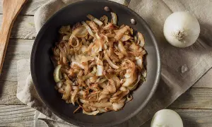 Gebratene Zwiebeln in einer Pfanne, goldbraun und karamellisiert. Nebenbei liegen zwei ganze Zwiebeln und ein Holzspatel. Perfekt zum Zwiebelgrillen!