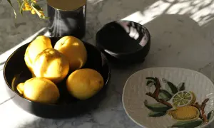 Frische, sonnengeküsste Zitronen lagern in einer schwarzen Schale auf Marmor. Daneben ein Teller mit Zitronenmotiv.