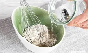 Eine Hand gießt Wasser aus einem Glas in eine Schüssel mit Roggenmehl und einem Schneebesen. Ein natürliches Shampoo aus Roggenmehl wird zubereitet.