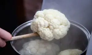 Ein Blumenkohl wird mit einem Holzlöffel aus kochendem Wasser gehoben. Das Blanchieren von Blumenkohl ist ein wichtiger Schritt.