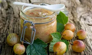 Glas mit selbstgemachter Stachelbeer Marmelade, umgeben von frischen Stachelbeeren und Blättern auf rustikalem Holz.