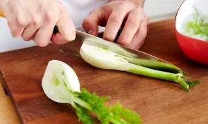 Hände schneiden frischen Fenchel für den Verzehr. Ein Teil liegt auf dem Holzbrett, ein anderer in einer roten Schüssel mit Wasser.