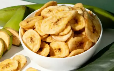 Schmackhafte bananenchips selber machen ist ganz einfach. Hier sind sie in einer Schüssel, daneben grüne Bananen und Blätter.