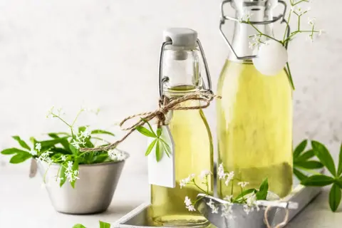 Zwei Flaschen mit hellem, gelbem Waldmeister Sirup, verziert mit frischen Waldmeisterzweigen und kleinen weißen Blüten.