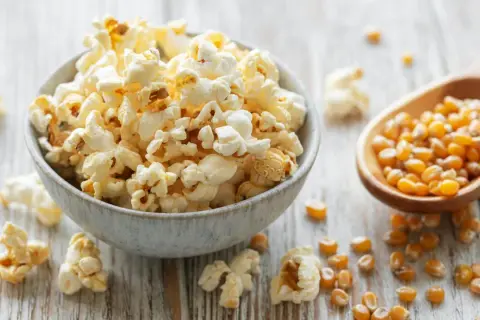 Schüssel mit frisch gemachtem Popcorn, daneben eine Holzschaufel mit Maiskörnern. So einfach ist Popcorn selber machen!