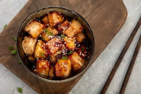 Ofen-gebackener Tofu in einer dunklen, glänzenden Sauce, bestreut mit Sesam und Frühlingszwiebeln, serviert in einer Schüssel auf einem Holzbrett.