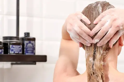Hände formen ein Herz in schaumigen Haaren. Pflegeprodukte stehen auf einem Regal im Hintergrund.