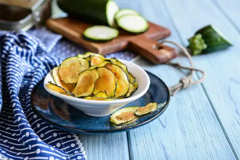 Knusprige Zucchini-Chips in einer Schüssel, daneben frische Zucchinischeiben auf einem Holzbrett.