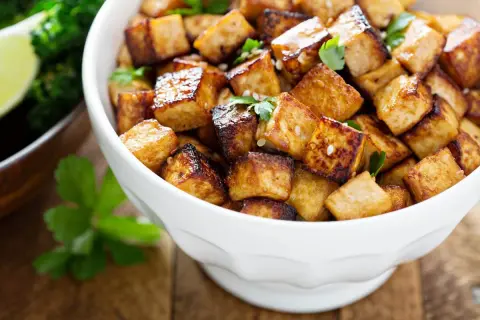 Knusprig gebratene Tofuwürfel, perfekt zum tofu zubereiten, garniert mit Petersilie und Sesam. Daneben ein Schälchen mit Brokkoli und Limette.