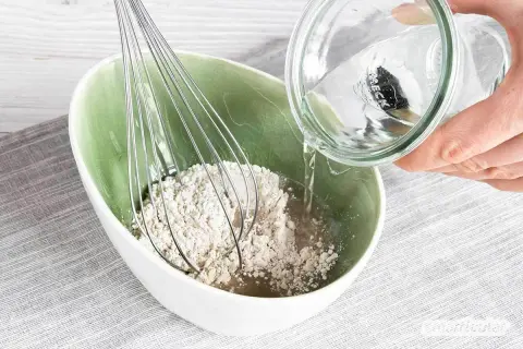 Eine Hand gießt Wasser aus einem Glas in eine Schüssel mit Roggenmehl und einem Schneebesen. Ein natürliches Shampoo aus Roggenmehl wird zubereitet.