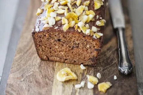 Saftiger bananenkuchen vegan mit Schokoladenglasur und Nüssen, auf einem Holzbrett mit einem Messer.