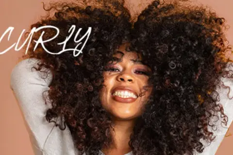 Frau mit vollem, lockigem Haar, die lächelt. Der Schriftzug "be CURLY" ist im Bild zu sehen.