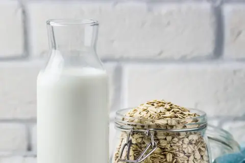 Glasflasche mit weißer Flüssigkeit neben einem Glas voller Haferflocken. Perfekt, um Hafermilch selber zu machen.