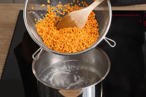 Rote Linsen werden aus einem Sieb in kochendes Wasser geschüttet. Das Kochen von Linsen ist einfach und schnell.