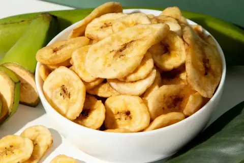 Schmackhafte bananenchips selber machen ist ganz einfach. Hier sind sie in einer Schüssel, daneben grüne Bananen und Blätter.
