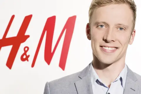 Junger Mann lächelt vor H&M-Logo. Kritische Stimmen werfen dem Unternehmen H&M Greenwashing vor, während es sich als nachhaltig präsentiert.