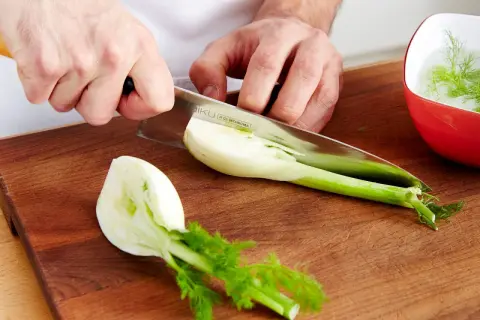Hände schneiden frischen Fenchel für den Verzehr. Ein Teil liegt auf dem Holzbrett, ein anderer in einer roten Schüssel mit Wasser.