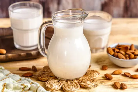 Frisch zubereitete Mandelmilch in einer Glaskaraffe, daneben Gläser und eine Schale mit Mandeln. So einfach ist Mandelmilch selber machen!