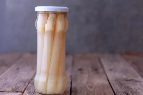 Ein Glas mit eingelegtem weißen Spargel steht auf einem Holztisch. Perfekt für das Spargel einkochen.