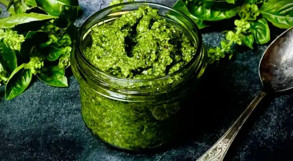 Ein Glas mit selbstgemachtem Pesto, umgeben von frischem Basilikum. Daneben liegt ein Löffel.