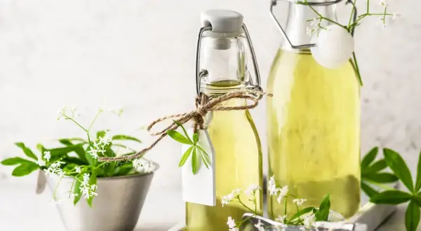 Zwei Flaschen mit hellem, gelbem Waldmeister Sirup, verziert mit frischen Waldmeisterzweigen und kleinen weißen Blüten.