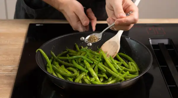 Frische brechbohnen kochen in der Pfanne. Gewürze werden mit einem Löffel hinzugefügt, während ein Kochlöffel die Bohnen wendet.