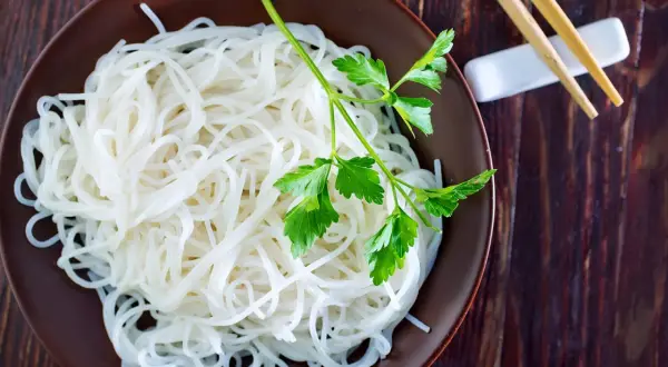 Reisnudeln kochen: Eine Schüssel voller zarter Reisnudeln, garniert mit frischer Petersilie, bereit zum Genießen. Stäbchen liegen daneben.