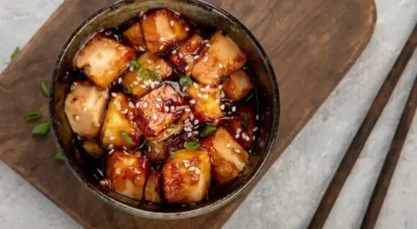 Ofen-gebackener Tofu in einer dunklen, glänzenden Sauce, bestreut mit Sesam und Frühlingszwiebeln, serviert in einer Schüssel auf einem Holzbrett.