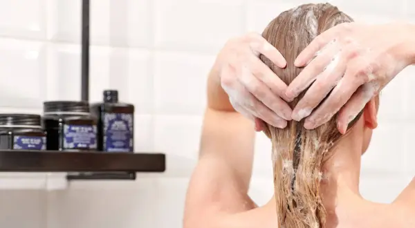 Hände formen ein Herz in schaumigen Haaren. Pflegeprodukte stehen auf einem Regal im Hintergrund.
