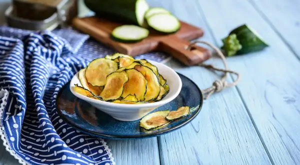 Knusprige Zucchini-Chips in einer Schüssel, daneben frische Zucchinischeiben auf einem Holzbrett.