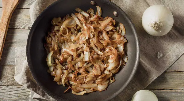 Gebratene Zwiebeln in einer Pfanne, goldbraun und karamellisiert. Nebenbei liegen zwei ganze Zwiebeln und ein Holzspatel. Perfekt zum Zwiebelgrillen!