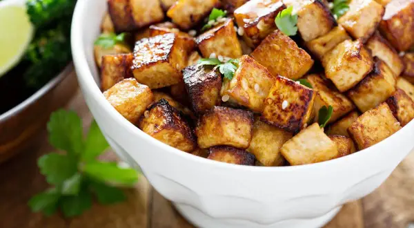 Knusprig gebratene Tofuwürfel, perfekt zum tofu zubereiten, garniert mit Petersilie und Sesam. Daneben ein Schälchen mit Brokkoli und Limette.