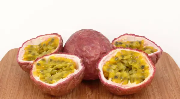 Ein ganzes und drei halbierte Passionsfrüchte liegen auf einem Holzbrett. Das Fruchtfleisch mit den Kernen lädt zum Passionsfrucht essen ein.