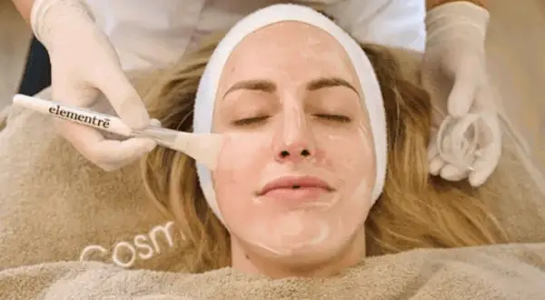 Eine Kosmetikerin trägt mit einem Pinsel eine Maske auf das Gesicht einer Frau auf, um es zu reinigen.