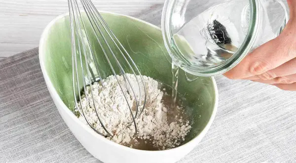 Eine Hand gießt Wasser aus einem Glas in eine Schüssel mit Roggenmehl und einem Schneebesen. Ein natürliches Shampoo aus Roggenmehl wird zubereitet.