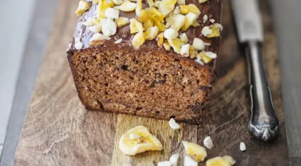 Saftiger bananenkuchen vegan mit Schokoladenglasur und Nüssen, auf einem Holzbrett mit einem Messer.