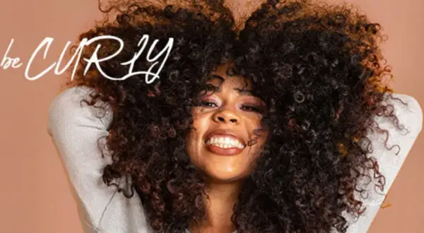 Frau mit vollem, lockigem Haar, die lächelt. Der Schriftzug "be CURLY" ist im Bild zu sehen.