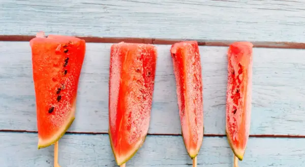 Vier gefrorene Wassermelonenstücke auf Holzstäbchen. Man kann Wassermelone einfrieren und so erfrischende Snacks für heiße Tage kreieren.