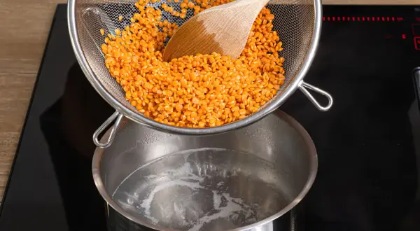 Rote Linsen werden aus einem Sieb in kochendes Wasser geschüttet. Das Kochen von Linsen ist einfach und schnell.