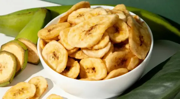 Schmackhafte bananenchips selber machen ist ganz einfach. Hier sind sie in einer Schüssel, daneben grüne Bananen und Blätter.