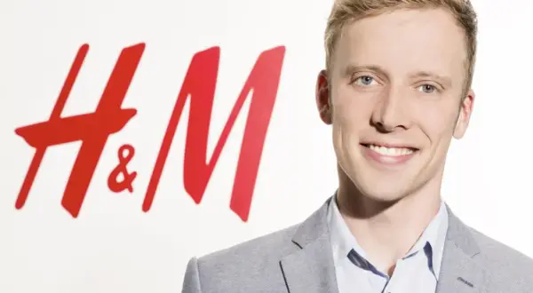 Junger Mann lächelt vor H&M-Logo. Kritische Stimmen werfen dem Unternehmen H&M Greenwashing vor, während es sich als nachhaltig präsentiert.