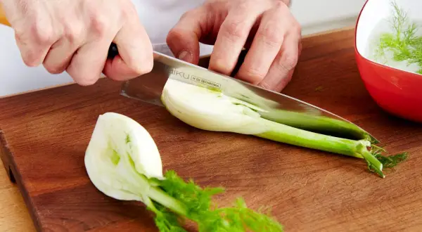 Hände schneiden frischen Fenchel für den Verzehr. Ein Teil liegt auf dem Holzbrett, ein anderer in einer roten Schüssel mit Wasser.