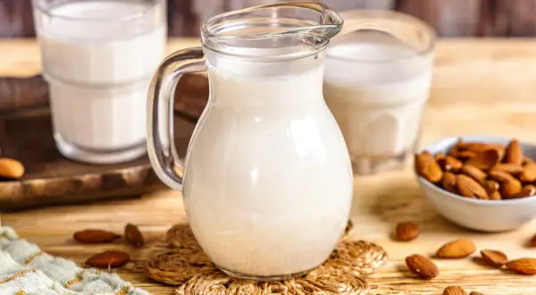 Frisch zubereitete Mandelmilch in einer Glaskaraffe, daneben Gläser und eine Schale mit Mandeln. So einfach ist Mandelmilch selber machen!