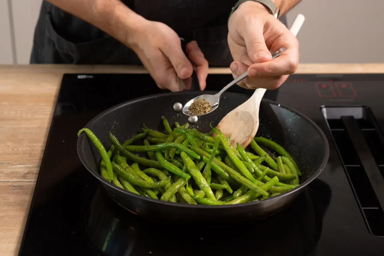 Frische brechbohnen kochen in der Pfanne. Gewürze werden mit einem Löffel hinzugefügt, während ein Kochlöffel die Bohnen wendet.