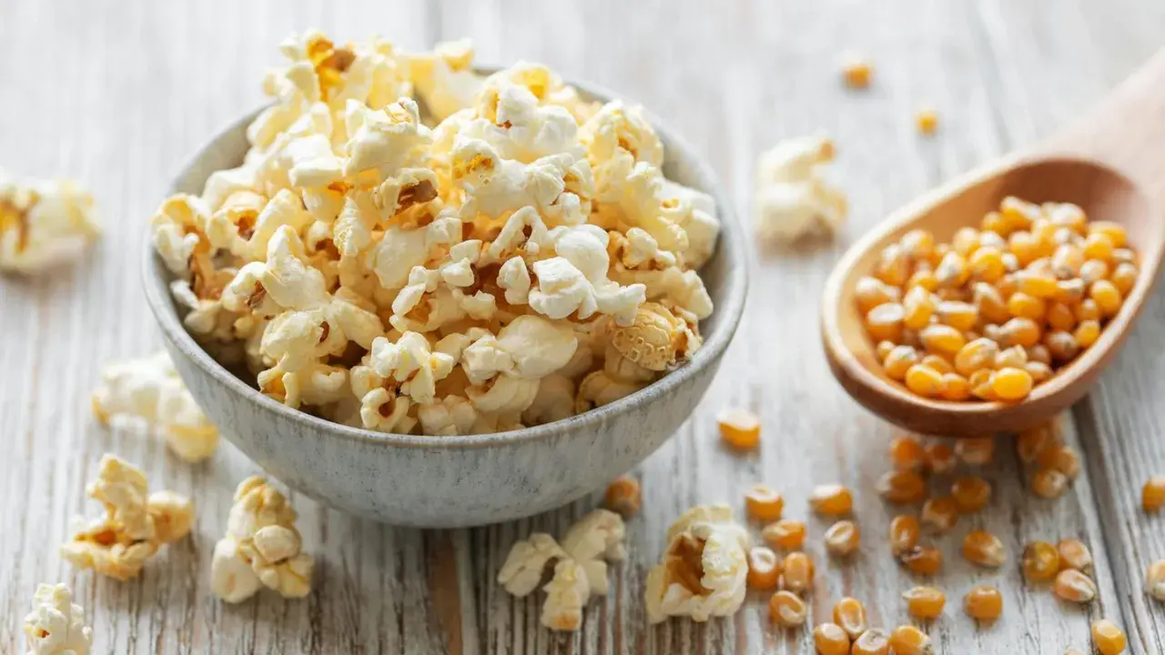 Schüssel mit frisch gemachtem Popcorn, daneben eine Holzschaufel mit Maiskörnern. So einfach ist Popcorn selber machen!