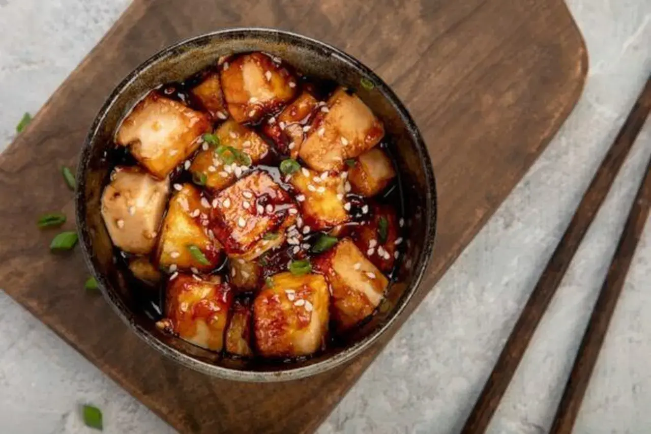 Ofen-gebackener Tofu in einer dunklen, glänzenden Sauce, bestreut mit Sesam und Frühlingszwiebeln, serviert in einer Schüssel auf einem Holzbrett.