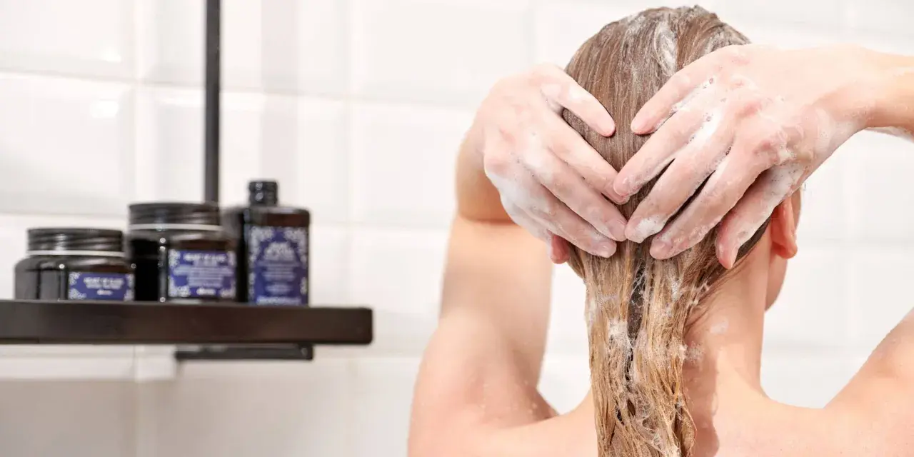 Hände formen ein Herz in schaumigen Haaren. Pflegeprodukte stehen auf einem Regal im Hintergrund.